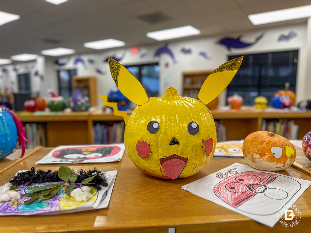 A bright yellow pumpkin decorated as Pikachu with paper ears, rosy cheeks, and a happy expression. Other decorated pumpkins and colorful student artwork surround it on the library shelf.