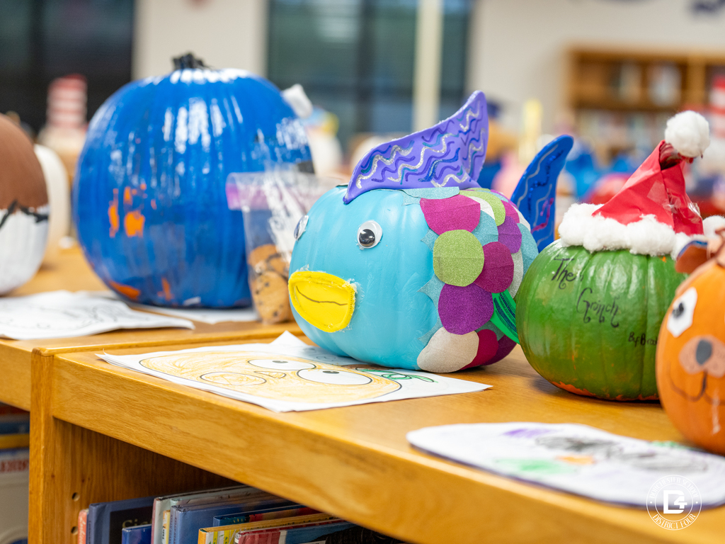 A blue pumpkin painted as the Rainbow Fish, with shiny, colorful fins and a bright yellow mouth. Next to it, a green pumpkin with a Santa hat is decorated as the Grinch.