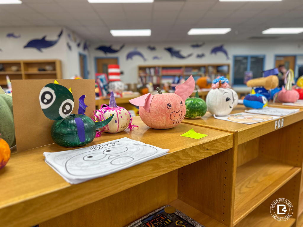 A lineup of decorated pumpkins on a library shelf, including a green dragon with paper wings and big cartoon eyes, a pink pig with paper ears, and other creative book-themed designs.