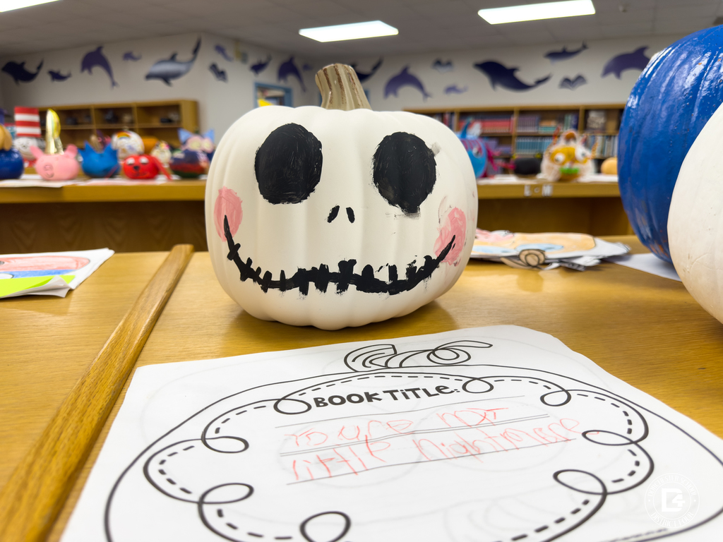 A white pumpkin painted with a large black smile, stitched mouth, and dark eyes resembling Jack Skellington from The Nightmare Before Christmas. It sits on a student’s worksheet labeled with the book title.