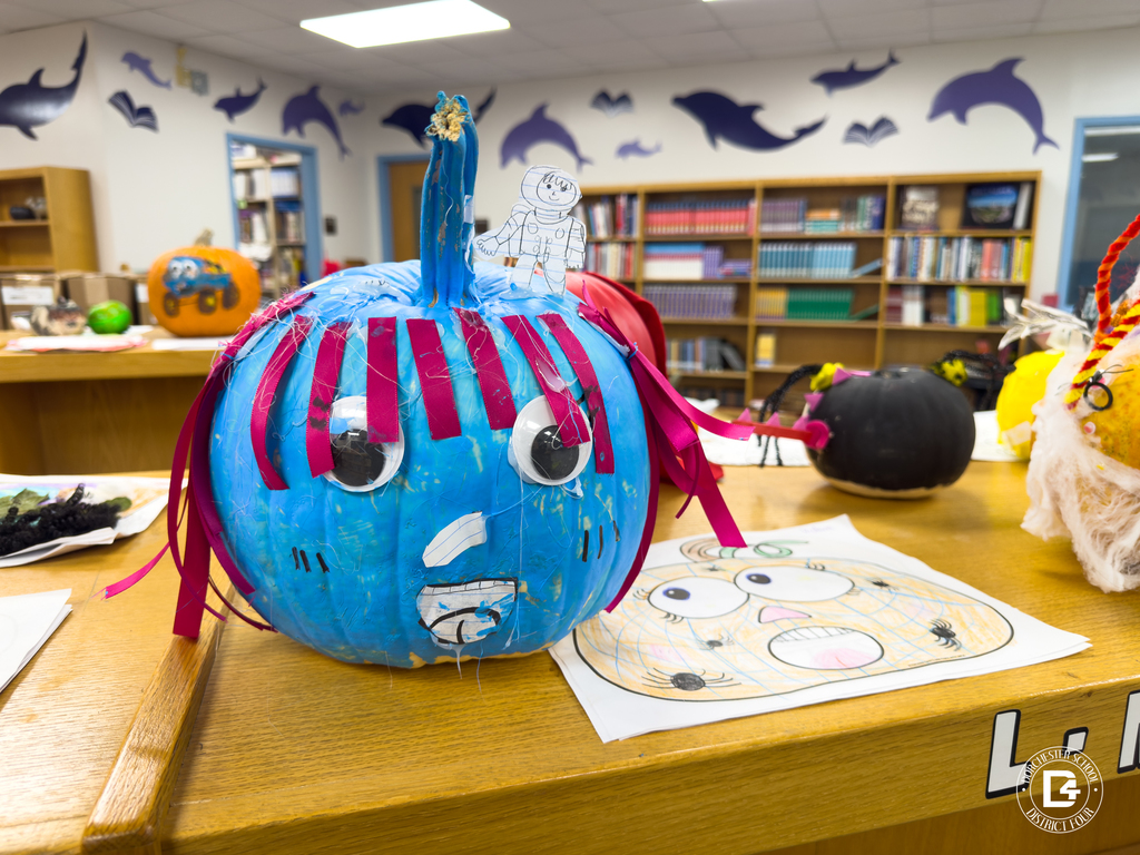 A blue pumpkin decorated as a storybook character with large white eyes, pink ribbon hair, and a paper astronaut figure taped to the stem. It sits on a table alongside a child’s pumpkin drawing.