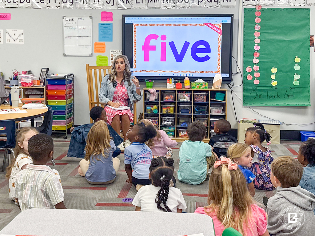 Mrs. Griffith leads a whole-group lesson from a rocking chair as students sit on the carpet, looking toward a screen displaying the word “five.”