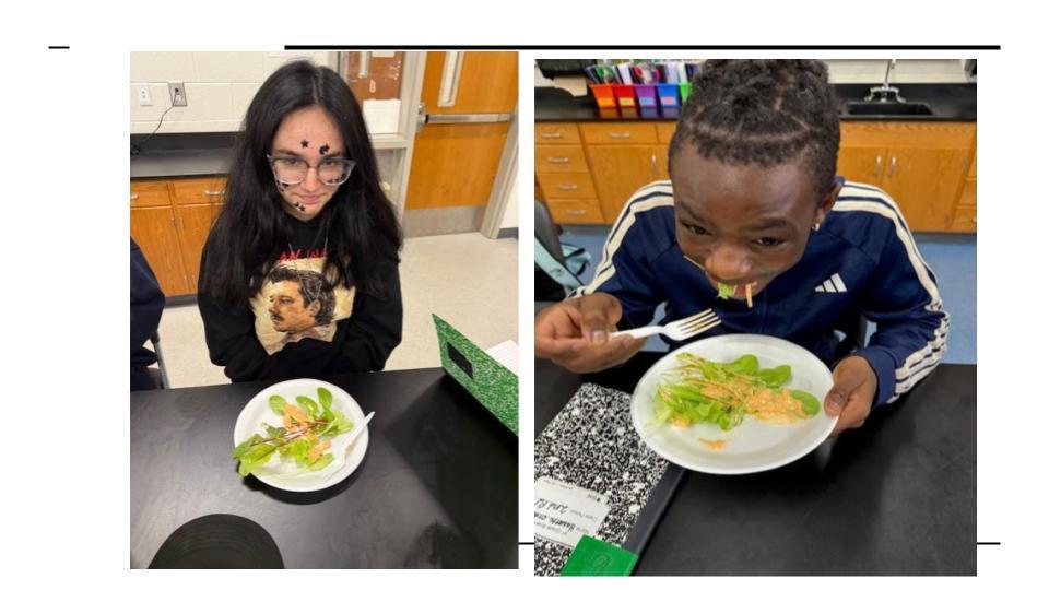 Two students sit at lab tables tasting fresh lettuce grown in class. On the left, a student wearing glasses and a black sweatshirt with a print design looks down at a small salad bowl. On the right, a student in a navy track jacket smiles while taking a big bite of lettuce using a fork.