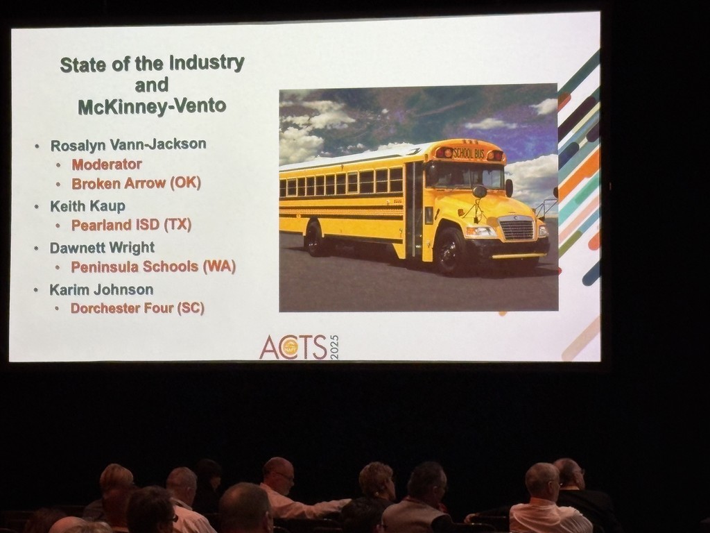 A presentation slide titled “State of the Industry and McKinney-Vento” is projected on a large screen. The slide lists speakers including Rosalyn Vann-Jackson (OK), Keith Kaup (TX), Dawnnett Wright (WA), and Karim Johnson (SC). A yellow school bus image appears beside the text, and attendees are seated in the audience below.