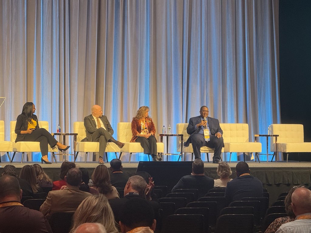 Four panelists sit on stage during a conference session with blue lighting and a beige curtain backdrop. Mr. Karim Johnson from Dorchester School District Four is speaking into a microphone while the other three panelists listen.