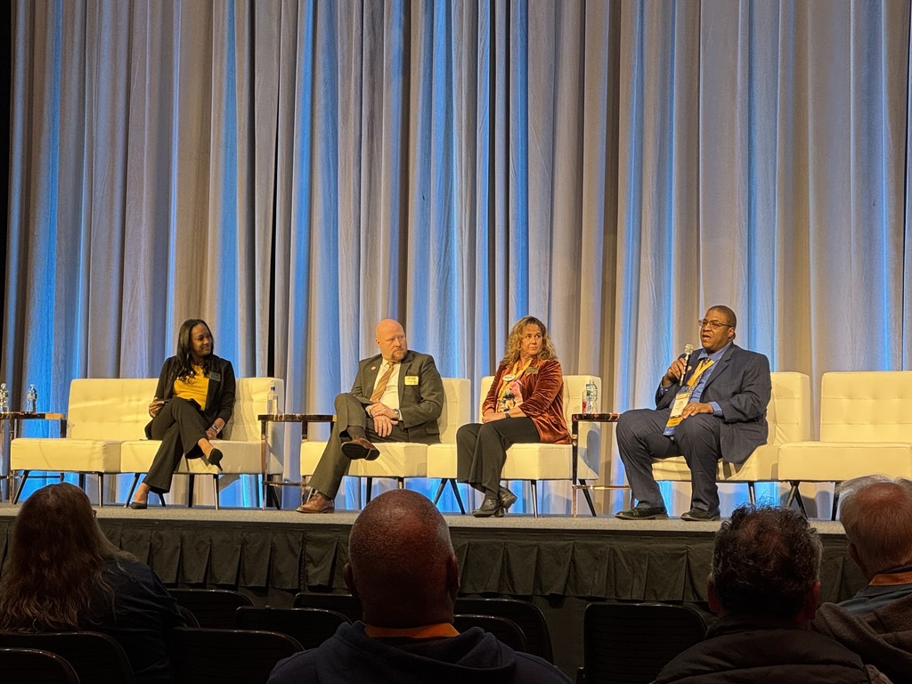 The same four panelists are seated on stage, engaging in a professional discussion. Mr. Karim Johnson, holding a microphone, speaks to the audience about McKinney-Vento Transportation.