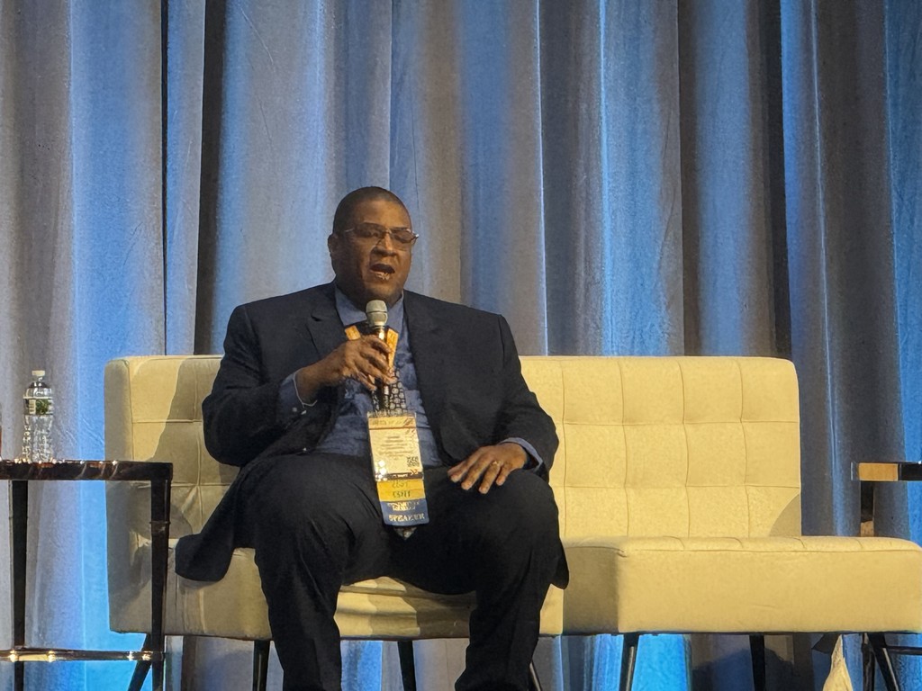 Mr. Karim Johnson, Director of Student Transportation for Dorchester School District Four, speaks confidently on stage into a microphone during the National Association for Pupil Transportation conference. A lanyard with multiple badges identifies him as a featured speaker.