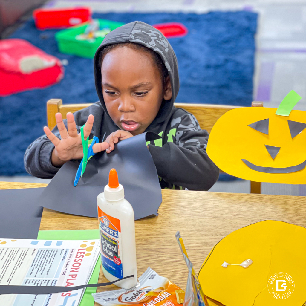 Parent and child working together at a table with paper, glue, and snacks while crafting pumpkins.