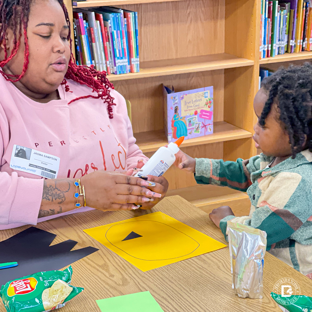 Parent and child working together at a table, gluing a black triangle shape onto a yellow pumpkin craft in a classroom library setting.