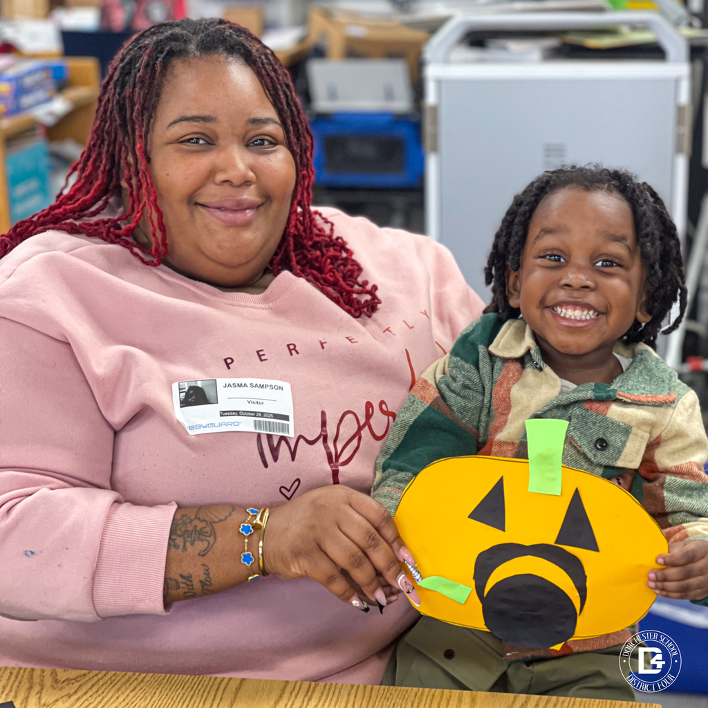 Parent and child sitting at a library table, smiling while showing their pumpkin craft with green and black shapes.