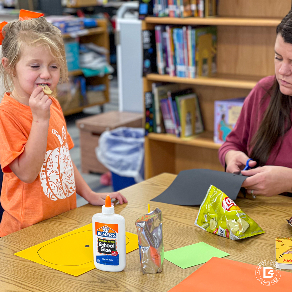 Child eating a snack beside their parent, who is cutting black paper for a pumpkin craft.