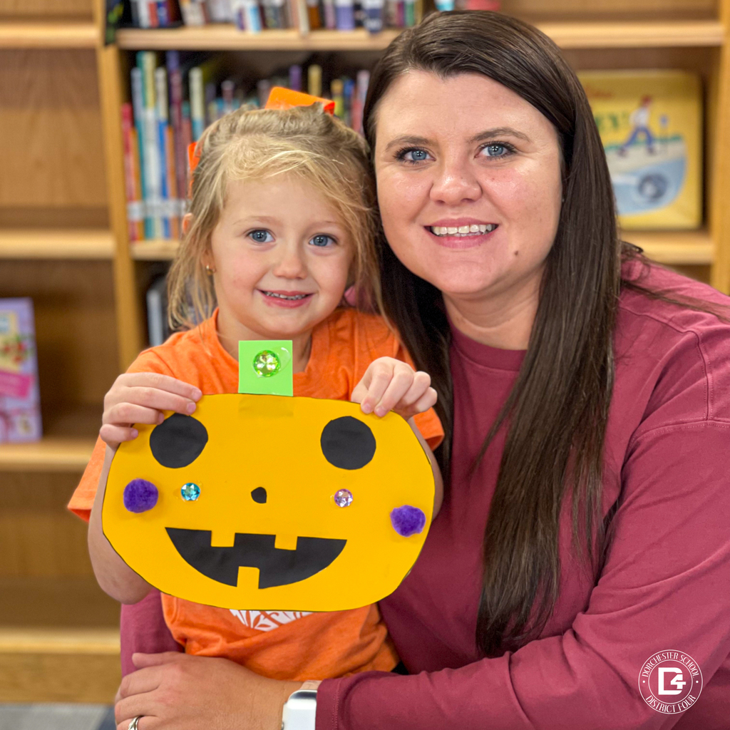 Parent and child smiling, holding a pumpkin craft with black cutout shapes and colorful decorations in a school library.