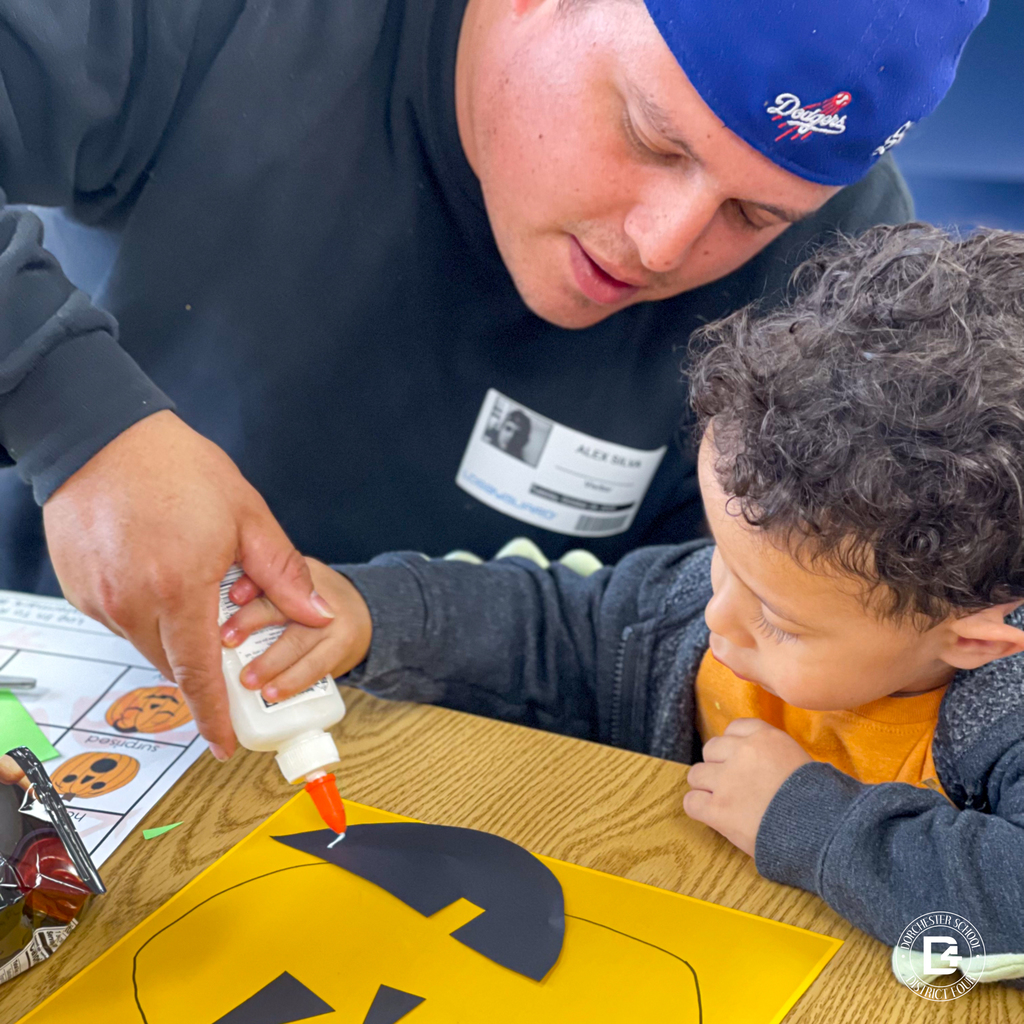Parent assisting a child with gluing shapes onto a yellow pumpkin cutout.