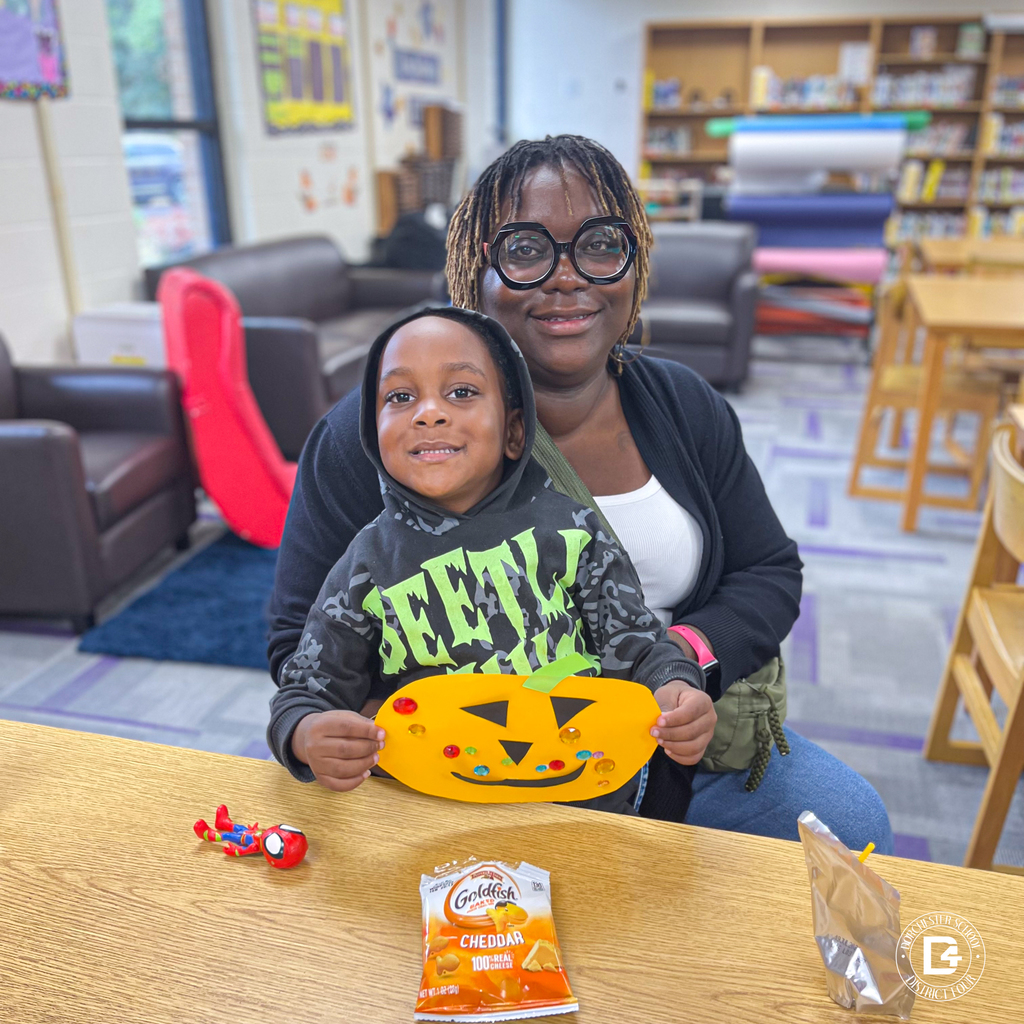 Parent and child smiling at a library table, holding a pumpkin craft decorated with gems and shapes; snacks and a juice pouch sit nearby.