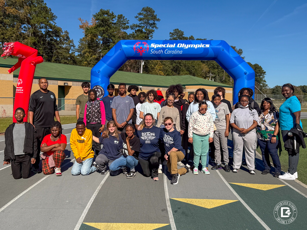 A large group of high school students and staff pose together on a track under a blue inflatable arch that reads “Special Olympics South Carolina.” A red air dancer waves nearby as the group smiles in the sunlight.