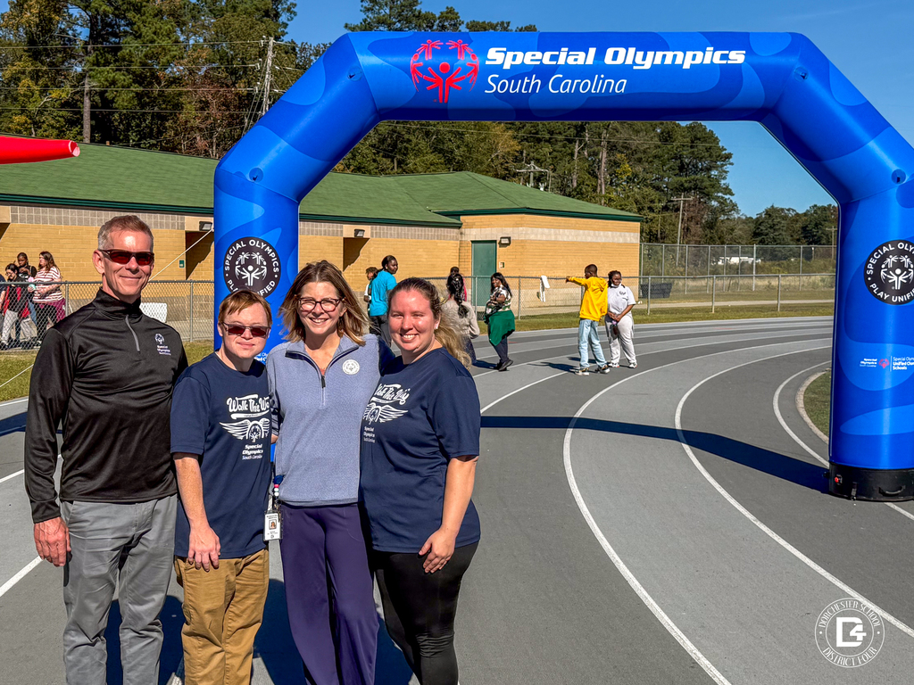 Four adults stand together under the same blue Special Olympics arch, smiling for a photo. They wear shirts with the event name “Walk the Way” while students and staff mingle in the background.