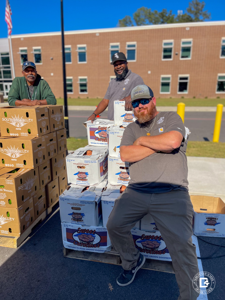 Three men from Dorchester School District Four pose beside stacks of produce boxes labeled “Sweet Carolinas” and “Southern Valley.” They stand in front of a brick school building under a clear blue sky, smiling as they help distribute food to families.