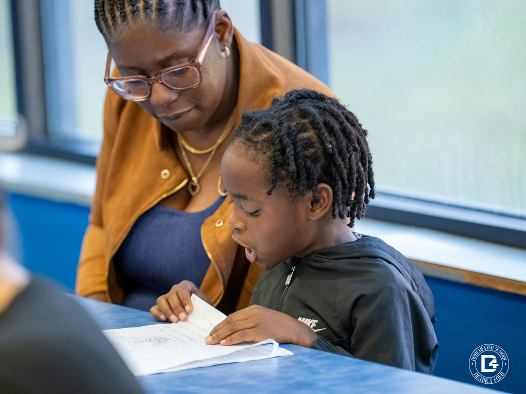 A boy shows his illustrated story to two adults seated across from him at a cafeteria table. The man and woman listen closely and smile as the student explains his work. Other families are gathered in the background.