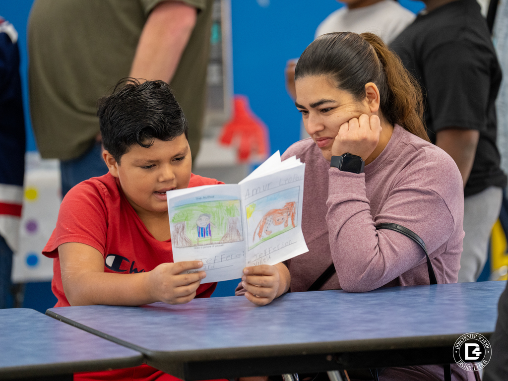 A young boy in a red shirt excitedly reads his handmade book to a woman beside him at a cafeteria table. The woman, wearing a pink sweater, smiles and listens closely as he shares his story.