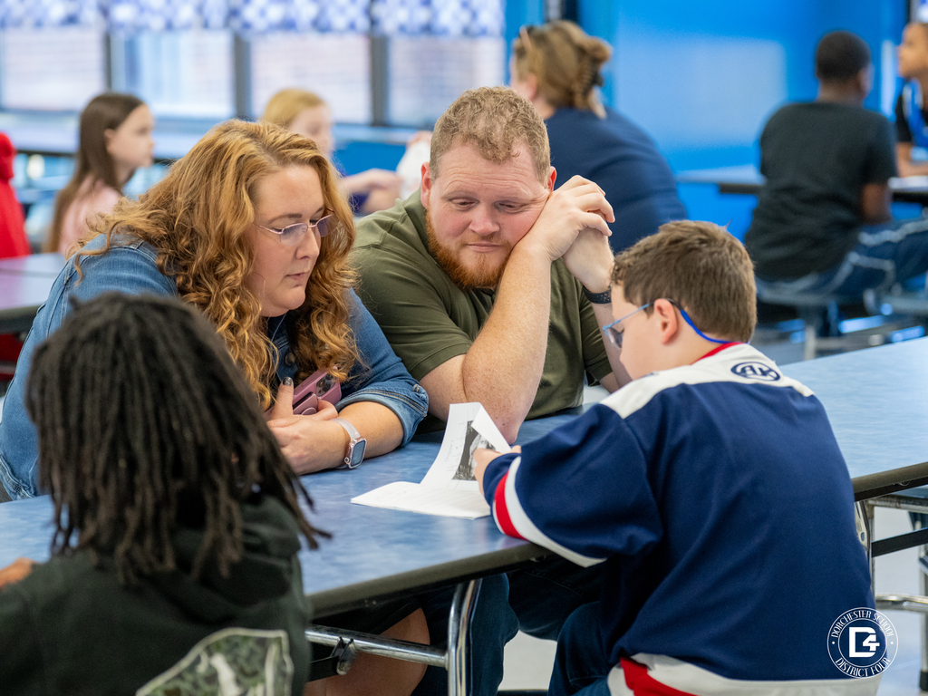 A young child reads their handwritten story to a woman beside them at a cafeteria table. The child’s expression is animated while reading, and the woman listens supportively, resting her hand on the table.