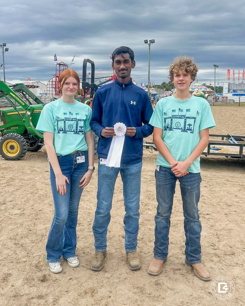 Three Woodland High School FFA students stand outdoors on a dirt lot at the Coastal Carolina Fair. The student in the middle holds a white ribbon, while the two others stand beside him smiling. They wear jeans and light blue FFA T-shirts, with fair rides and equipment visible in the background under a cloudy sky.