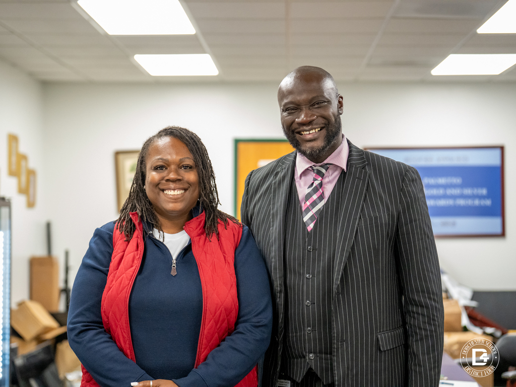 Mrs. Bryant and Mr. White pose in an office, smiling together. Mr. White wears a pinstriped suit, and Mrs. Bryant wears a red vest over a navy shirt.