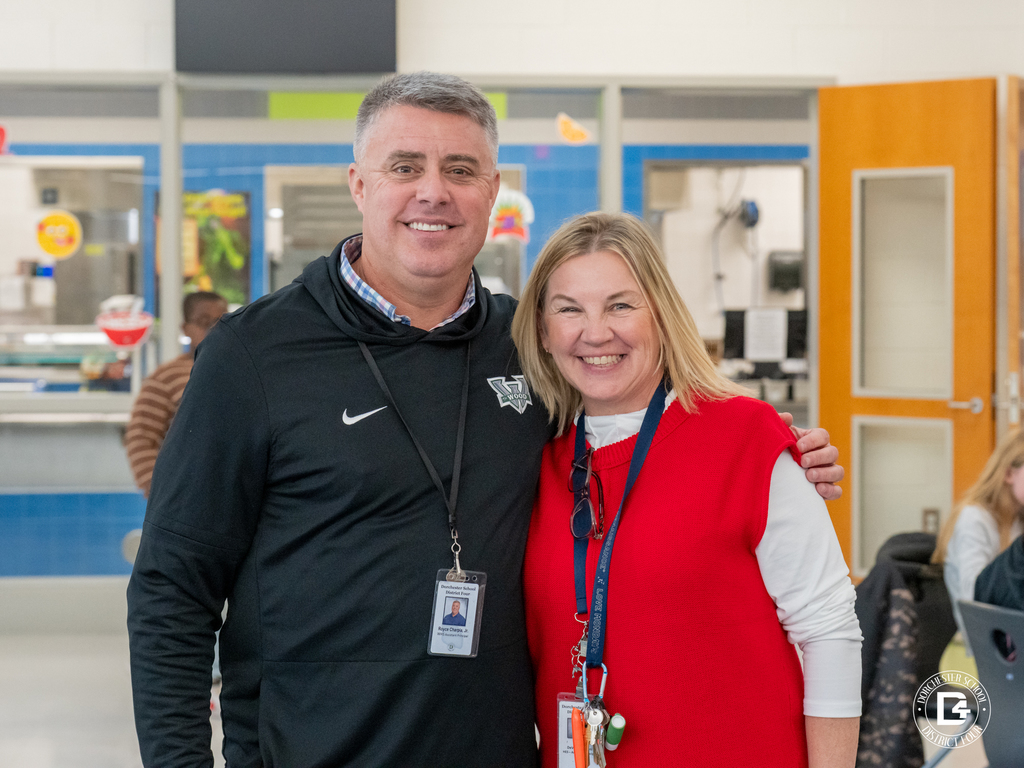 Mr. Charpia and Mrs. Trigiani stand side by side in a cafeteria, smiling warmly at the camera. Both wear ID badges and casual professional attire.