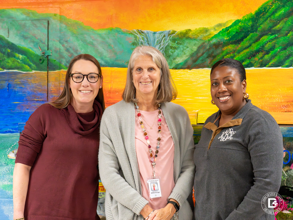 Mrs. Hartzog, Ms. Mims and Mrs. Busby pose in front of a brightly painted mural featuring a landscape scene. All three smile warmly at the camera.