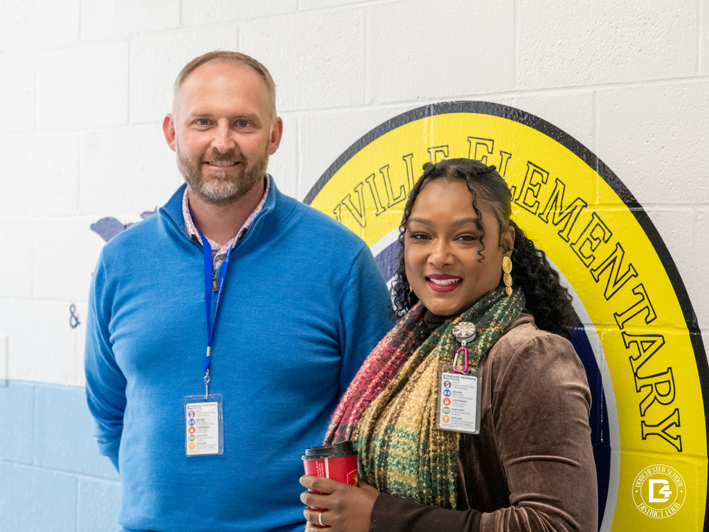 Mr. Wolfgang and Mrs. Butler stand in front of a school wall featuring a colorful circular emblem. They smile at the camera, both wearing professional attire and lanyards.