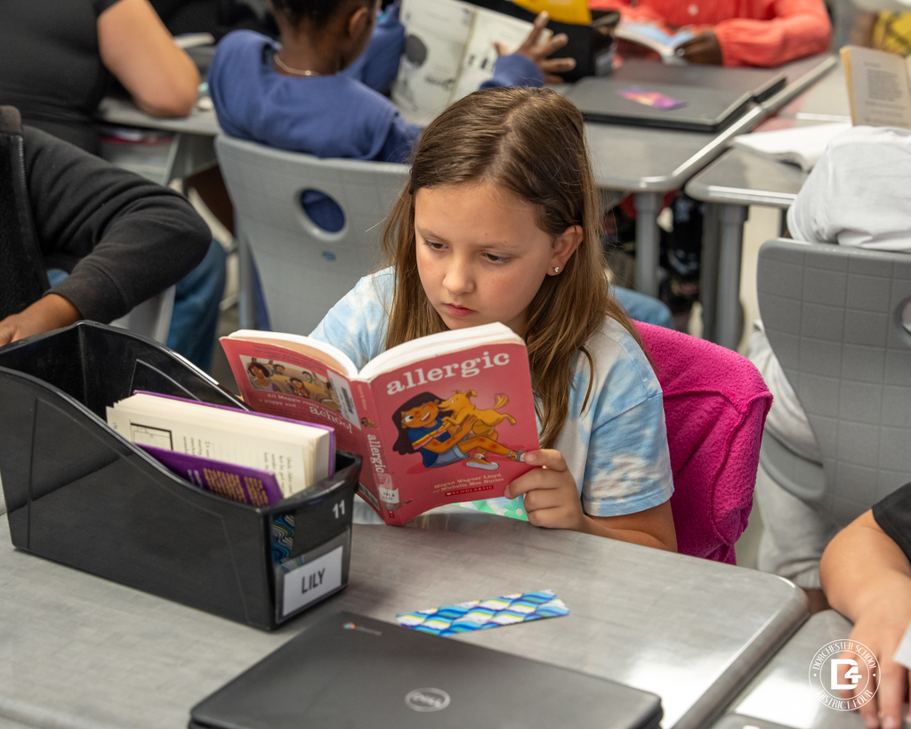 A student in a tie-dye shirt reads a chapter book titled “Allergic” while seated at a classroom table with other students reading nearby.