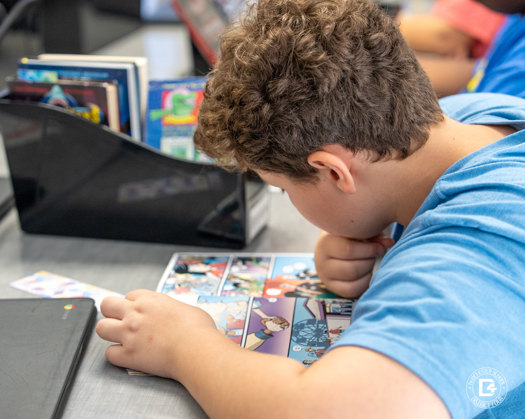 A 5th-grade student wearing a blue shirt reads a colorful comic book at his desk, surrounded by books and a Chromebook.