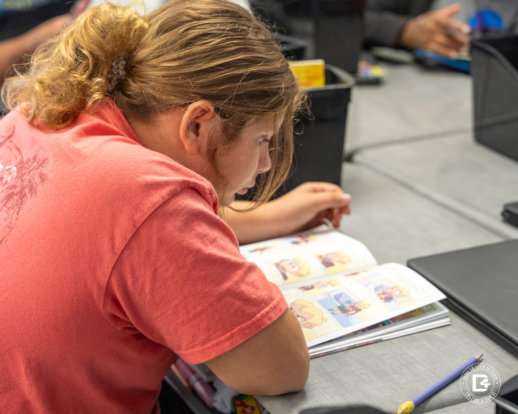 A student in a red shirt leans over her desk, focused on reading an illustrated book with a pencil and Chromebook nearby.