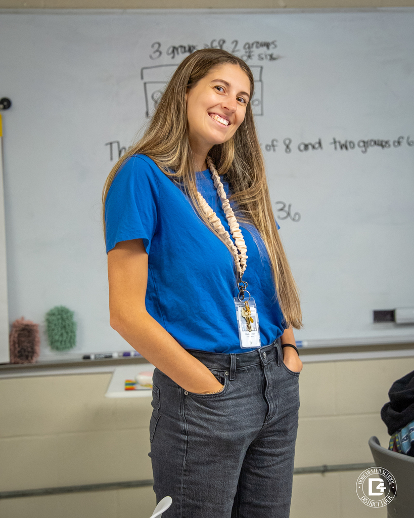 Ms. Huff, wearing a blue shirt and gray pants, smiles warmly in front of her classroom whiteboard, ready to inspire her 5th graders.