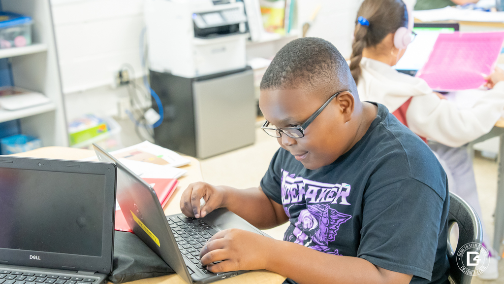 A student in a black T-shirt types on a Chromebook, concentrating on an online math activity.