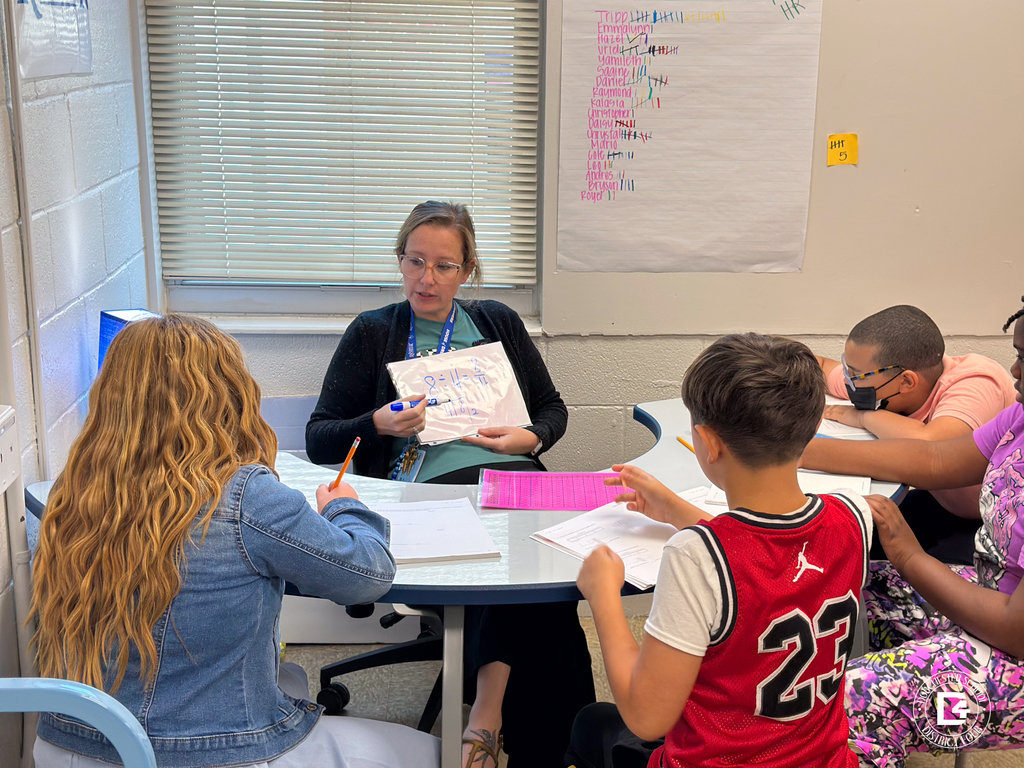 Mrs. Pafford leads a small group at a round table, holding up a whiteboard with a math equation as students follow along.