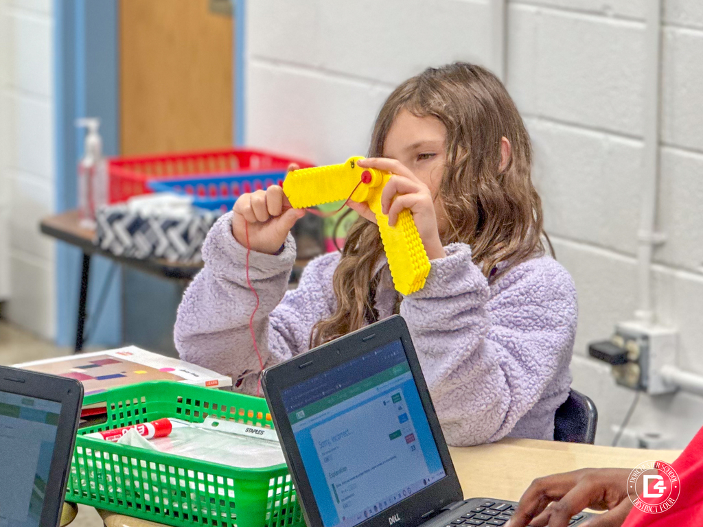 A student in a lavender fleece jacket threads red yarn through a yellow pegboard as part of a hands-on math activity.