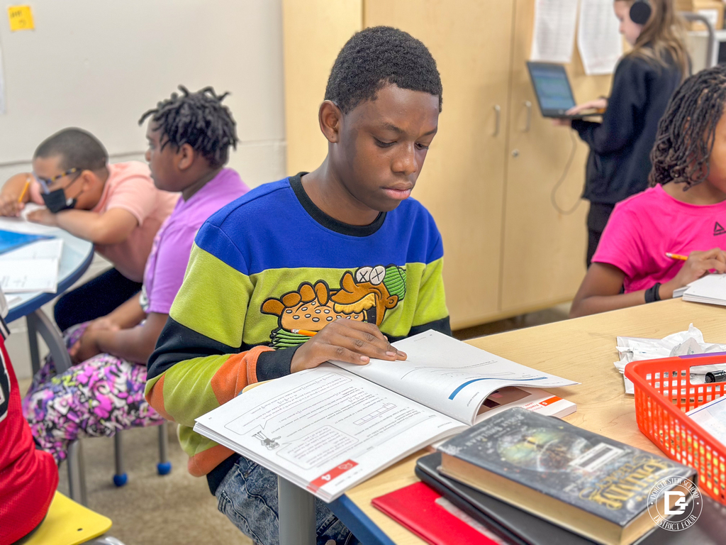 A student wearing a colorful sweater reads and completes a math workbook activity while seated at a table.