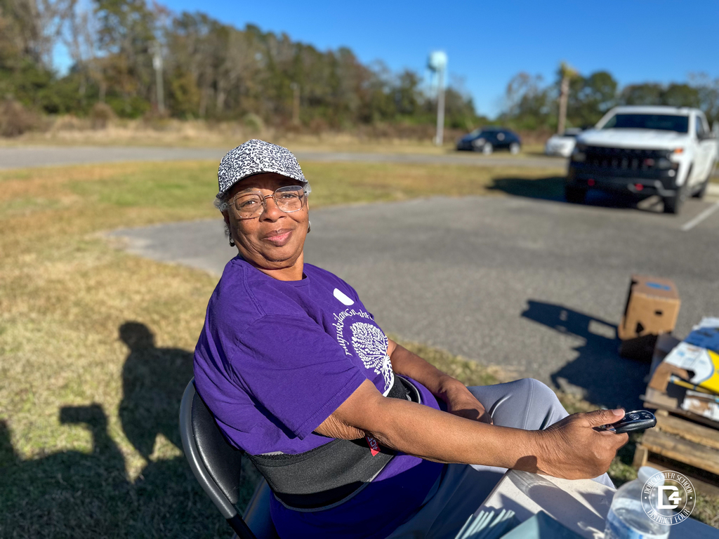A woman sitting in a folding chair wearing a purple shirt and patterned hat, smiling while holding a cell phone.