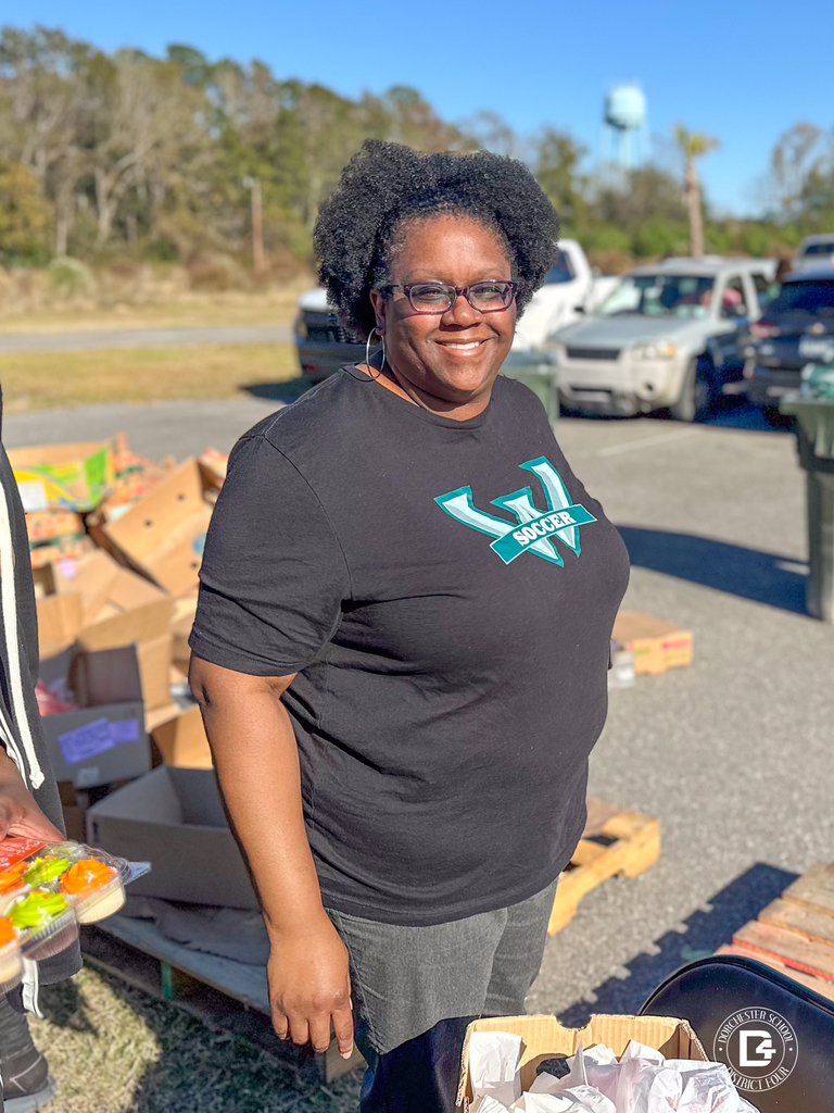 A woman wearing a black shirt with a V Soccer logo, smiling while standing beside boxes of distributed food.
