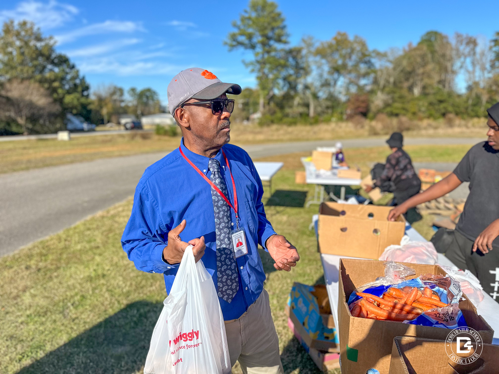 A man wearing a blue shirt, tie, and Clemson hat holding a grocery bag while standing near boxes of produce at the distribution site.