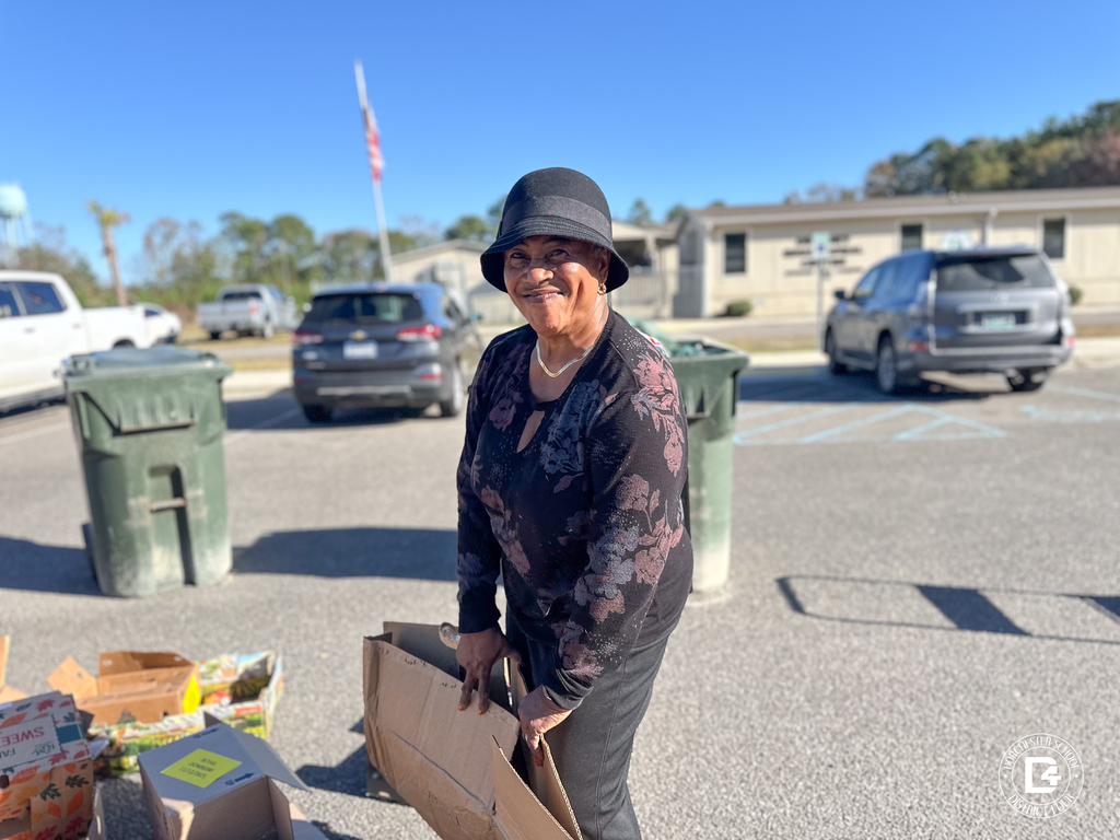 A woman wearing a black hat and patterned top bending down to break down cardboard boxes in the parking lot.