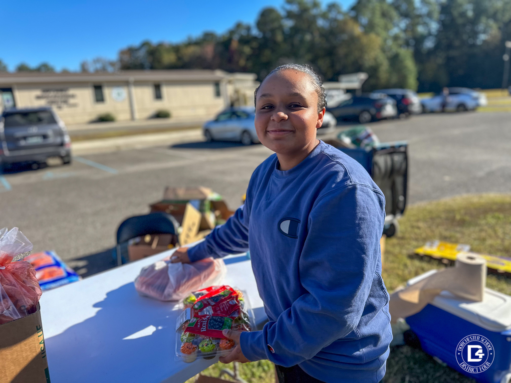 A student wearing a blue sweatshirt smiling at the camera while helping pack items at a distribution table.