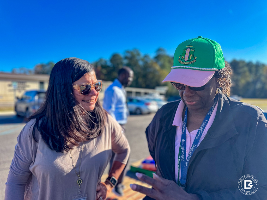 Two women talking and smiling outdoors, one wearing sunglasses and the other wearing a green and pink cap and district lanyard.
