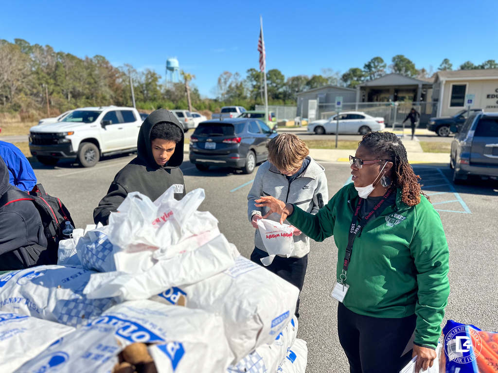 A group of students and a staff member sorting and bagging food in a parking lot with cars and school buildings in the background.