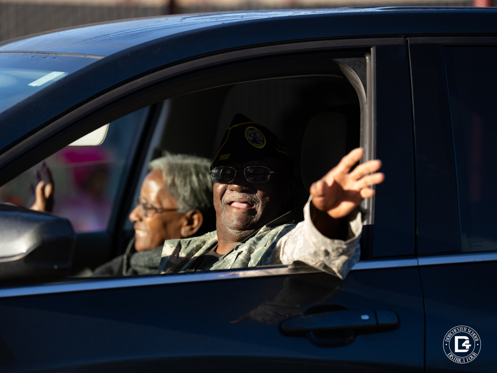 A veteran in uniform waves from a car window, smiling, with another passenger seated beside him.