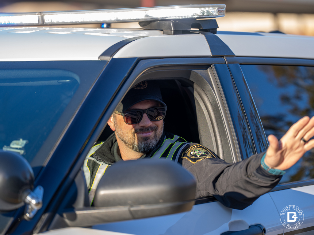 A uniformed officer drives by in a patrol vehicle, smiling and waving out the window.