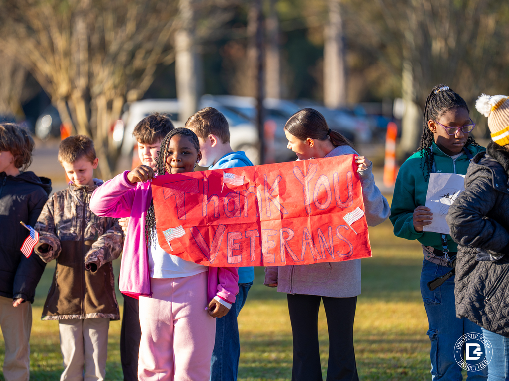 Students stand outside holding a large red sign that reads Thank You Veterans while others nearby wave small American flags.