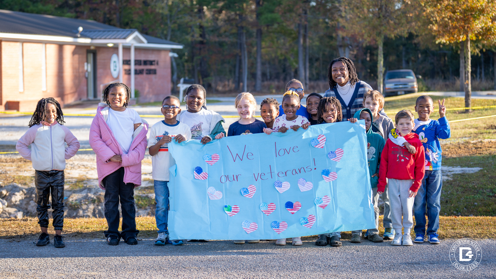 A class of students stands together holding a large blue banner reading We love our veterans decorated with American flag-themed paper hearts.