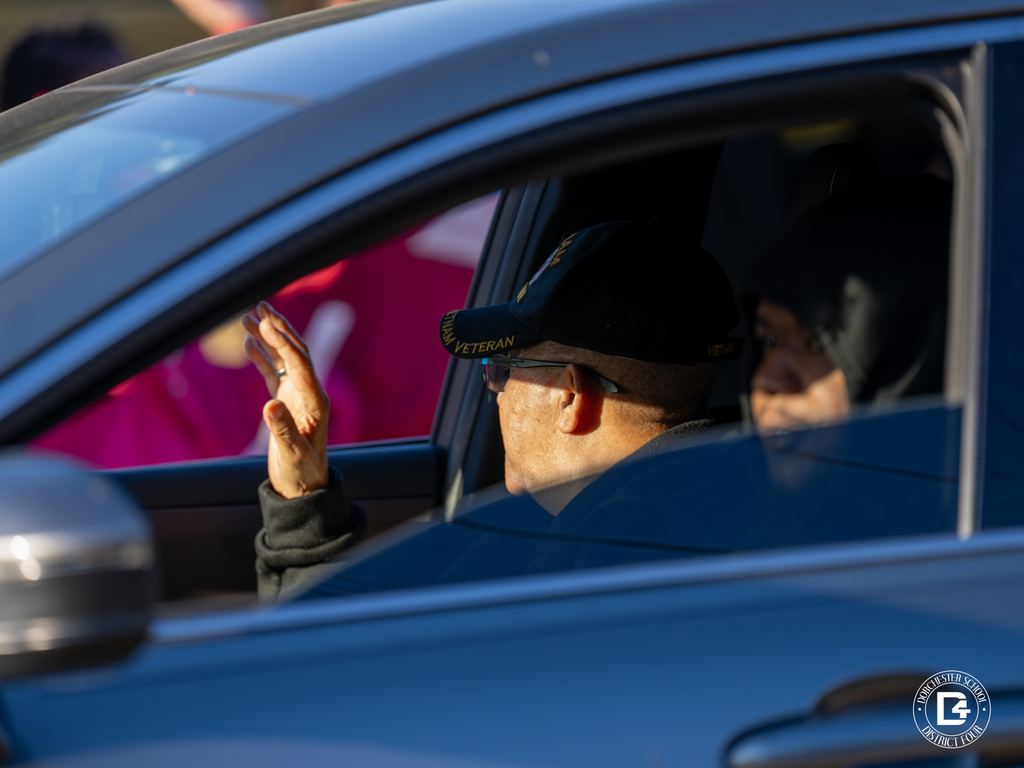 A veteran wearing a service cap waves out the window of a car while a passenger sits beside him.