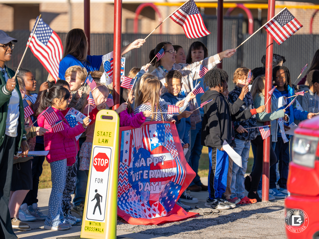 A large group of students waves American flags and cheers as parade vehicles pass through the car rider loop.